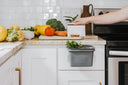 Person using a small gray compost bin in a kitchen with fruits and vegetables on the counter.