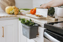 Kitchen counter with vegetables, a cutting board, and a gray container for food waste.